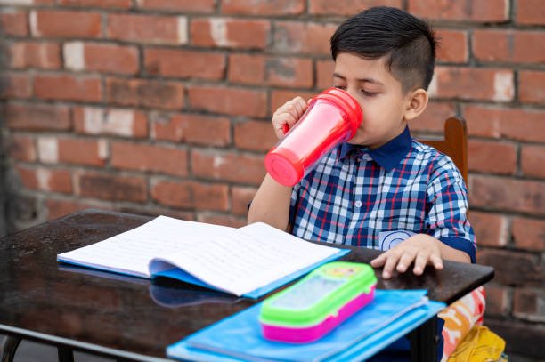 Primary school student drinking water with red water bottle while sitting in class on study table. Indian school student in classroom.